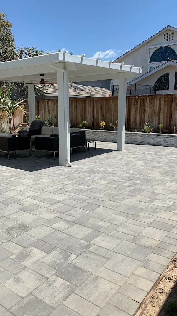 White pergola over gray paver patio with planter beds and wooden fence